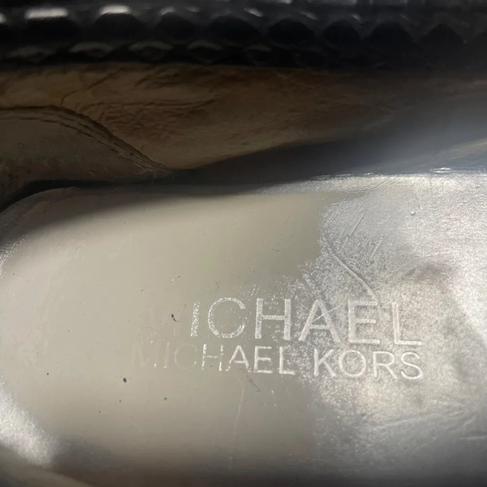 Snygga svarta ballerinaskor från Michael Kors med krokodilpräglat mönster och en stor silverfärgad MK-logga på framsidan. Skorna har en elegant och stilren design, perfekt för både vardag och fest. De är i bra skick med lite slitage på insidan.. Skor.