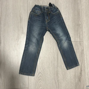 Barnjeans från Denim Day - Säljer ett par snygga barnjeans från Denim Day i storlek 104. De är i en klassisk blå färg med slitna detaljer och har en elastisk midja för extra komfort. Perfekta för vardagsbruk och lek!