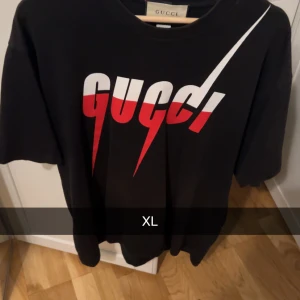 Svart t-shirt från Gucci - Säljer en snygg svart t-shirt från Gucci i storlek XL. Den har ett coolt tryck i vitt och rött på framsidan. Perfekt för en avslappnad och stilren look. Passar bra till både jeans och shorts. Perfekt för sommaren eller våren!