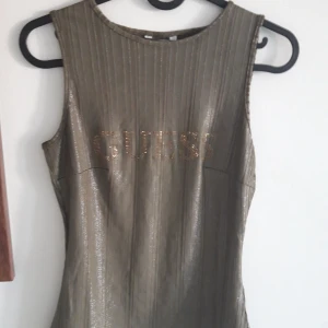 Glittrig topp från Guess - Snygg glittrig topp från Guess (strl S) i en metallisk grön-brun nyans. Den är ärmlös och har ett ribbat mönster med Guess-loggan i glittriga stenar på framsidan. Perfekt för en festlig kväll eller en utekväll med vänner. Toppen är stretchig och framhäver figuren.