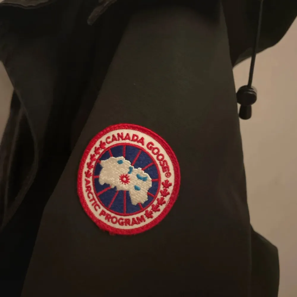 Säljer en svart parkajacka från Canada Goose. Jackan har en klassisk design med huva och flera praktiska fickor. Perfekt för kyliga höst- och vinterdagar. Den har en snygg broderad logga på ärmen. Passar både till vardags och mer äventyrliga utflykter. Jackan har små skador som Inte syns i verkligheten men hör av er för bilder/frågor. Jackor.