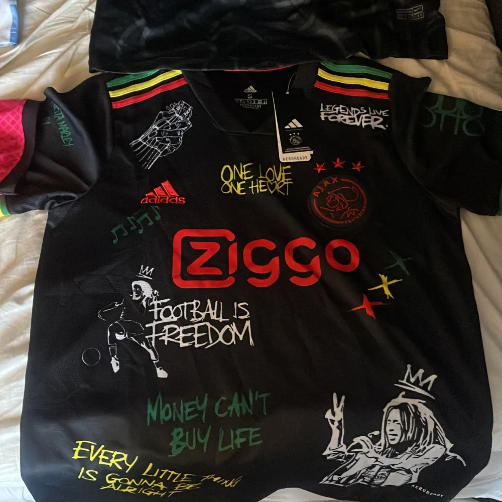 Säljer en unik svart Ajax fotbollströja från Adidas med Bob Marley-tema. Tröjan har coola detaljer som färgglada ränder på axlarna och text som 'One Love, One Heart'. Den är kortärmad och i nytt skick. Perfekt för fotbollsfans som älskar reggae och stil! 🌟. T-shirts.