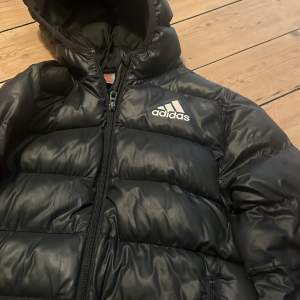 Säljer en svart dunjacka från Adidas i storlek 9-10 år. Jackan har en dragkedja och en mysig huva, perfekt för kalla dagar. Den är i bra skick men har en liten reva som syns på bilden. Perfekt för vintern! ❄️