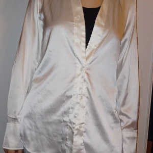 Vit satinblus - Säljer en elegant vit satinblus med långa ärmar och knappar framtill. Perfekt för både fest och vardag. Blusen har en v-ringad design som ger en stilren look. Passar bra till både jeans och kjol. Skicka ett meddelande vid intresse!