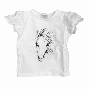 Vit t-shirt med hästmotiv - Vit t-shirt med hästmotiv på framsidan. Den har korta ärmar med volangdetaljer och knappar vid axeln för enkel påklädning.