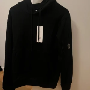 Svart hoodie från C.P. Company - Säljer en svart hoodie från C.P. Company i nytt skick. Den har en klassisk design med en justerbar huva och en ficka framtill. Perfekt för både vardag och chill. Kommer i originalförpackning. Bastant M