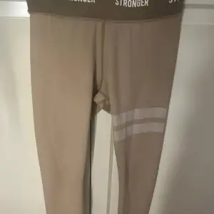 Säljer ett par snygga beige leggings från Stronger i storlek S. De har en hög midja med elastiskt band där det står 'STRONGER'. Perfekta för träning eller en avslappnad dag. De har en diskret randig detalj på ena benet. Superbekväma och stilrena!