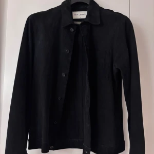 Svart mockajacka från Our Legacy - Säljer en stilren svart mockajacka från Our Legacy. Jackan har en klassisk krage och knappar framtill. Perfekt för höst och vår, med en normal passform och långärmade ärmar. Den är i mycket bra skick och passar både till vardags och fest.