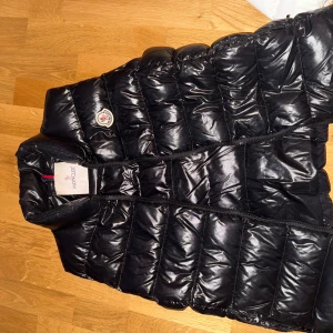 Svart dunväst från Moncler - Säljer en snygg svart dunväst från Moncler i mycket bra skick. Västen har en glansig finish och är perfekt för höst och vinter. Den har en dragkedja framtill och Monclers ikoniska logga på bröstet. Perfekt för att hålla värmen med stil! Finns kvitto digitalt från Monclers hemsida, samt orginalförpackning.
