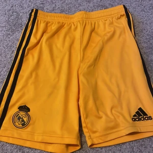 Gula fotbollsshorts från Adidas - Säljer ett par gula fotbollsshorts från Adidas med Real Madrid-logga. De har svarta ränder på sidorna och är i bra skick. Perfekta för fotbollsträningen eller att bara chilla i. Storleken är inte angiven, men de ser ut att vara i en standard herrstorlek.