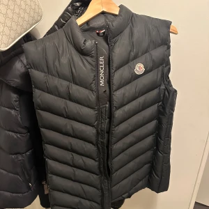 Svart dunväst från Moncler - Säljer en snygg svart dunväst från Moncler i mycket bra skick. Västen har en dragkedja framtill och Monclers logga på bröstet. Perfekt för höst och vinter, håller dig varm och stilren. Passar både till vardags och lite finare tillfällen.