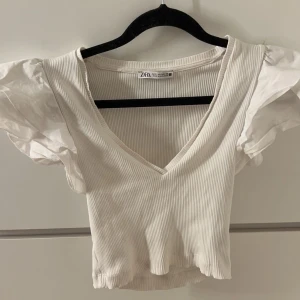 Vit ribbad topp från Zara - Säljer en superfin vit ribbad topp från Zara. Den har en v-ringad design och puffiga kortärmar Perfekt för sommardagar eller fest. Toppen har inga deferenser💞