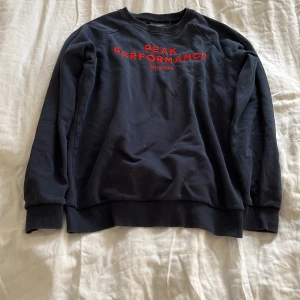 Marinblå sweatshirt från Peak Performance - Säljer en marinblå sweatshirt från Peak Performance med röd broderad text på framsidan. Tröjan har långa ärmar och ribbade muddar vid ärmslut och nederkant. Perfekt för en avslappnad stil eller som ett extra lager under kalla dagar.