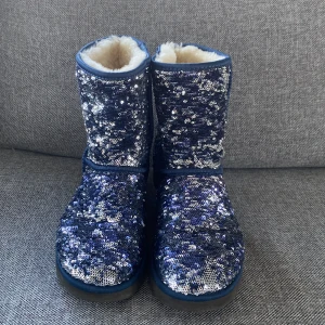 Glittriga blå UGG boots - Säljer ett par supercoola UGG boots i blått med glittriga paljetter över hela ytan. De är fodrade med mjukt och varmt foder, perfekt för kalla dagar. Sulan är robust och ger bra grepp. Dessa boots är både snygga och bekväma, idealiska för vinterns alla äventyr! ✨