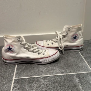 Vita Converse All Star - Säljer ett par klassiska vita Converse All Star i bra skick. De har den ikoniska höga modellen med snörning och röd/blå rand runt sulan. Perfekta för både vardag och fest! Lite slitna men fortfarande snygga och bekväma.