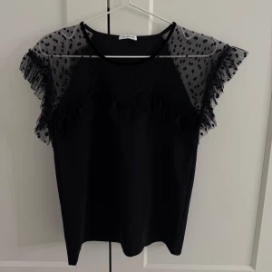 Ida Sjöstedt topp  - Snygg svart blus från Ida Sjöstedt med genomskinliga spetsdetaljer och volangärmar. Perfekt för en festlig kväll eller en stilren vardagslook. Blusen har en elegant passform och är tillverkad i ett mjukt material som känns skönt mot huden.