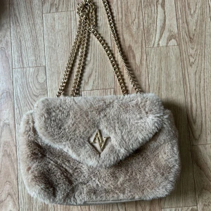 Beige pälsväska med kedja - Superfin beige axelväska i pälsmaterial med guldfärgad kedja. Väskan har en snygg logga på framsidan och är perfekt för att ge en lyxig touch till din outfit. Den är mjuk och fluffig, vilket gör den både trendig och mysig. Perfekt för höst och vinter! Inga defekter alls knappt använd💕