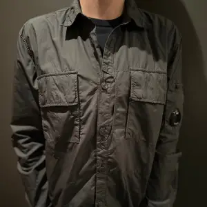 CP company Overshirt/Skjorta i grå/grön färg. Perfekt skick och utrustad med CP Companys ikoniska lins detalj på ärmen ger den en riktigt fräsch look. Den är perfekt som en lätt jacka eller ett extra lager och är designad för både stil och funktion. Nypris runt 4000kr⚡️