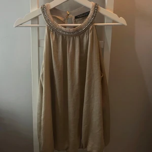 Beige ärmlös topp från Dorothy Perkins - Snygg beige ärmlös topp från Dorothy Perkins i storlek M. Den har en glittrig detalj runt halsringningen.