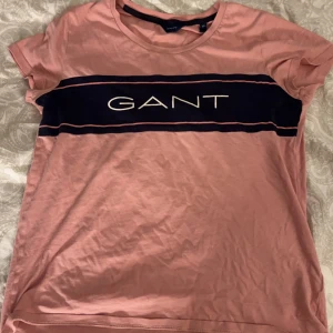 Rosa t-shirt från GANT - Säljer en snygg rosa t-shirt från GANT i storlek XS. Den har en mörkblå rand över bröstet med GANT-loggan i vitt. Perfekt för en avslappnad stil eller vardagsbruk. T-shirten är kortärmad och i bra skick.