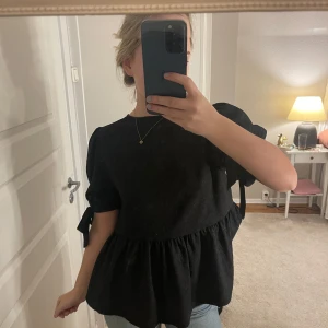 Svart blus med volangdetalj - Säljer en svart blus i mycket bra skick. Den har korta ärmar med knytband och en volangdetalj vid midjan som ger en fin siluett. Perfekt för både vardag och fest! Storlek s💕
