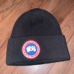  Canada Goose mössa - Säljer en svart ribbad mössa från Canada Goose i nytt skick. Den har en broderad logga framtill och är perfekt för kalla vinterdagar. Mössan är både stilren och varm, vilket gör den till ett måste i garderoben. Passar perfekt för vinterns alla äventyr! 🖤