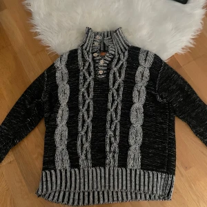 Svart och grå stickad tröja från Guan To - Säljer en snygg svart och grå stickad tröja från Guan To i storlek XL. Tröjan har ett flätat mönster framtill och ribbade detaljer vid krage, ärmslut och nederkant. Perfekt för kyliga dagar och ger en mysig känsla. Passar både till vardags och lite finare tillfällen. 