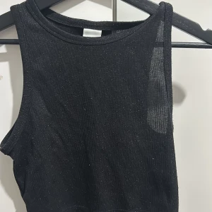 Svart glittrig ärmlös topp från H&M - Snygg svart ärmlös topp från H&M med glittrande detaljer. Perfekt för en utekväll eller fest! Toppen är ribbad och har en tight passform som framhäver figuren. Ett måste i garderoben för alla som älskar att glittra!