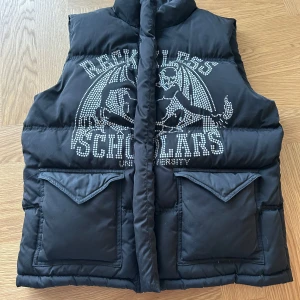 Reckless scholars dunväst - Snygg svart dunväst från Reckless Scholars med ett coolt tryck på framsidan och text på baksidan. Västen har en dragkedja framtill och två stora fickor med lock. Perfekt för att hålla sig varm och stilren.