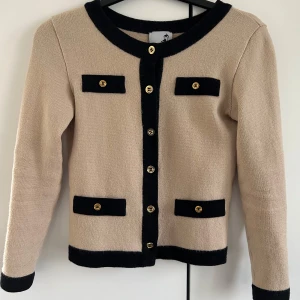 Beige kofta med svarta detaljer - Elegant beige kofta med svarta kanter och guldknappar. Den har fyra dekorativa fickor framtill och långa ärmar med knappar vid ärmsluten. Perfekt för en stilren look.