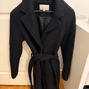 Svart kappa från Vila - Snygg svart kappa från Vila med knytbälte i midjan och knappar framtill. Den har en klassisk krage och långa ärmar, perfekt för kyligare dagar. Fodrad insida för extra komfort. En stilren och tidlös design som funkar till många tillfällen.