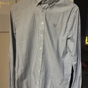Randig skjorta från GANT - Snygg randig skjorta från GANT i blått och vitt. Skjortan har långa ärmar och knappar framtill. Perfekt för en stilren look.