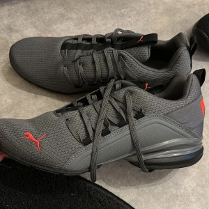 Grå sneakers från Puma - Snygga grå sneakers från Puma med röda detaljer och snörning. Skorna har en modern design med SoftFoam för extra komfort. Perfekta för både träning och vardag.