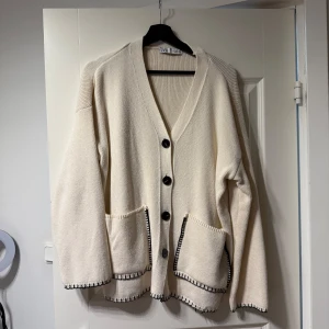 Beige kofta med svarta knappar - Säljer en stilren beige kofta från Zara med svarta knappar och två stora fickor framtill. Koftan har långa ärmar och en ribbad kantdetalj vid ärmslut och nederkant. Perfekt för en avslappnad look.