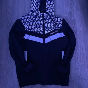 Svart hoodie från Black Squad - Säljer en svart hoodie från Black Squad med mönstrad huva och axelparti. Hoodien har dragkedja framtill och vita detaljer på bröstet. Perfekt för en casual look.