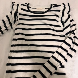 Randig tröja med volangdetaljer - Säljer en stilren randig tröja med svarta och vita ränder. Tröjan har långa ärmar och fina volangdetaljer vid axlarna. Perfekt för en trendig look!