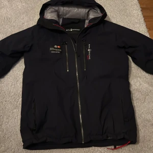 Svart jacka från Sail Racing - Snygg svart jacka från Sail Racing med huva och dragkedja. Jackan har en stilren design med logotyp på bröstet och ryggen. Insidan är grå och quiltad för extra värme. Perfekt för kyligare dagar. Pris kan diskuteras 