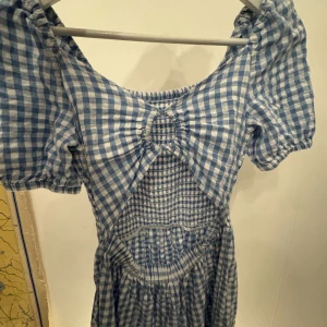 Blå och vit rutig klänning - Charmig blå och vit rutig klänning med puffiga kortärmar och en söt öppen ryggdetalj. Perfekt för sommarens äventyr med sin luftiga design och lekfulla mönster. Klänningen har en smickrande passform och en knytning i ryggen som ger extra stilpoäng.