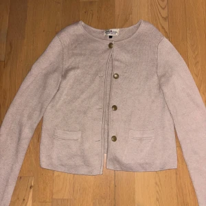 Beige kofta från Hampton Republic - Säljer en stilren beige kofta från Hampton Republic. Den har en rund halsringning och knäpps med fem dekorativa knappar framtill. Koftan är tillverkad i en mjuk blandning av bomull, viskos och nylon, vilket gör den både bekväm och hållbar. Perfekt för en avslappnad look.