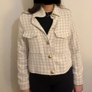 Beige rutig jacka med guldknappar - Snygg beige jacka med rutigt mönster och stiliga guldknappar. Jackan har en klassisk krage och två bröstfickor med lock. Perfekt för att ge en elegant touch till din outfit. Köpt från Zara