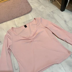 Rosa ribbad topp - Säljer en söt rosa ribbad topp med trekvartsärm och v-ringad design. Perfekt för en avslappnad stil. Toppen har en fin detalj vid halsringningen som ger en extra touch.