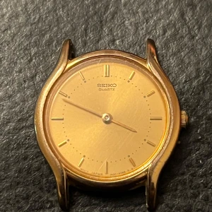 Guldfärgad Seiko Quartz-klocka - Elegant guldfärgad Seiko Quartz-klocka med minimalistisk design. Klockan har en rund urtavla med guldtonade visare och index. Baksidan är i rostfritt stål med ingraverad information. Perfekt för en stilren look.