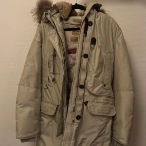 Beige parkajacka från Parajumpers - Säljer en snygg beige parkajacka från Parajumpers med pälskantad huva och flera praktiska fickor. Jackan har både dragkedja och knappar framtill samt en justerbar midja. Perfekt för kalla dagar! För fler information är det bara att skriva! Priset kan diskuteras! ÄKTA!