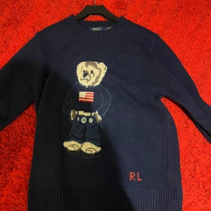 Polo Ralph Lauren - Säljer en mörkblå stickad tröja från Polo Ralph Lauren med en söt björn på framsidan. Tröjan har långa ärmar och en ribbad kant vid halsen. Perfekt för en avslappnad och stilren look.