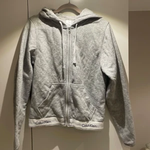 Grå hoodie från Calvin Klein - Säljer en stilren grå hoodie från Calvin Klein med dragkedja och huva. Tröjan har långa ärmar och praktiska fickor framtill. Calvin Klein-loggan syns på nederkanten. Perfekt för en avslappnad look.
