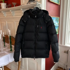 Svart dunjacka från Ralph Lauren Gorham - Svart dunjacka från Ralph Lauren Polo med röd logga på bröstet. Jackan var en dragkedja framtill, en bröstficka, två sidfickor samt en innerficka. Jackan har justerbara snören vid fållen så du kan vika upp den, går att justera bra. Äkta Dun! Har även orderbekräftelse. Säljer då den inte kommer till användning. Med Vänliga Hälsningar Thor!