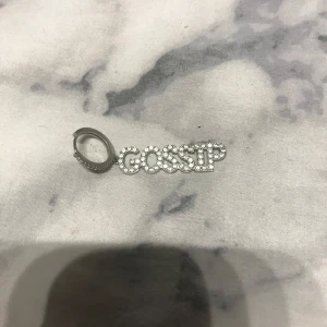 Silverfärgat örhänge med texten 'GOSSIP' - Stilrent och trendigt örhänge i silverfärg med texten 'GOSSIP' dekorerat med glittrande stenar. Perfekt för att ge en extra touch till din outfit.