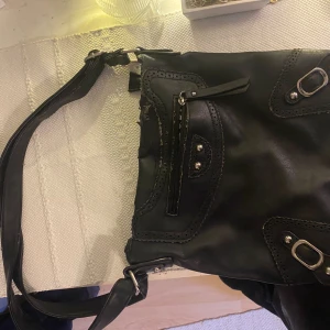 Svart axelväska med mönstrat foder - Snygg svart axelväska i skinn med dekorativa nitar och spännen. Väskan har ett mönstrat foder och en praktisk dragkedja för säker förvaring. Perfekt för att bära med sig det viktigaste med stil.