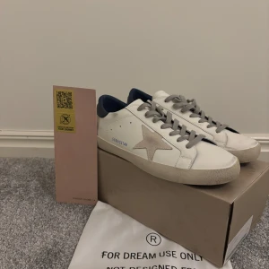 Golden Goose  - Snygga vita sneakers från Golden Goose med en ikonisk stjärndetalj på sidan. Skorna har grå snörning och en mörkblå häl. Perfekta för en stilren look.