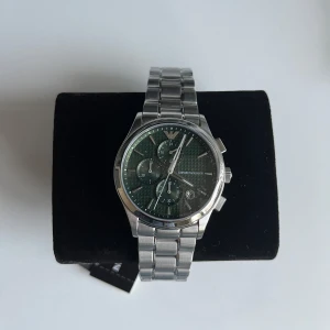  klocka Emporio Armani - Stilren klocka från Emporio Armani med silverfärgad länkarmband och mörk urtavla. Klockan har flera sub-dials och en elegant design som passar perfekt för den modeintresserade. Levereras med originalförpackning och certifikat. Oanvänd och köpt för 3700 kr, ligger fortfarande i plasten!
